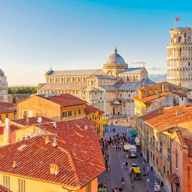 Aerial view of Pisa showing the Leaning Tower, Cathedral, and Baptistery with red-tiled rooftops. Visitors walk along the street below under blue sky.