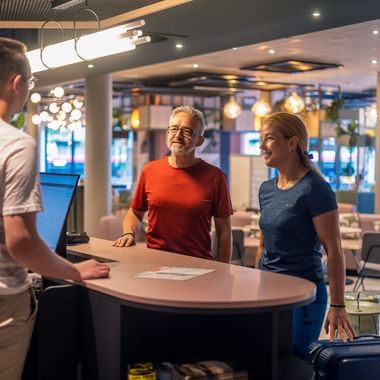 Lächelndes Paar mit Gepäck an der Hotelrezeption im Gespräch mit Mitarbeiter. Moderne Lobby mit Sitzbereich und großen Fenstern im Hintergrund.