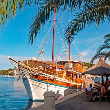 Traditionelles Holzsegelschiff MS Orkan an einer Uferpromenade festgemacht. Palmen rahmen die Szene, mit Café-Terrasse und blauem Himmel.