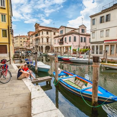 Two cyclists with bikes rest by a canal in Chioggia. Colorful boats are moored along wooden posts. Historic buildings line the waterfront under blue sky.