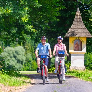 Two cyclists on red bikes riding past a traditional Austrian wayside shrine with yellow facade on a tree-lined path in Upper Austria.