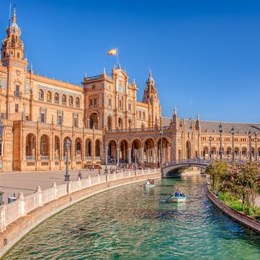 Plaza de España in Sevilla mit verzierten Backsteingebäuden, Arkadengang und Kanal mit Brücke unter klarem blauen Himmel.
