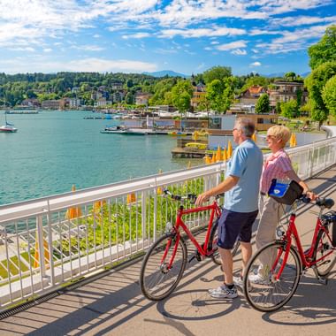 Two cyclists with red bikes pause at a lakeside railing in Velden am Wörthersee, overlooking turquoise water, boats, and the town of Velden.