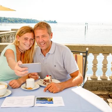 Lächelndes Paar macht Selfie auf Seeterrasse in Konstanz mit Kaffee und Kuchen, mit Blick auf den Bodensee mit Balustrade und Sonnenschirmen.
