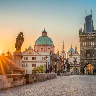 Karlsbrücke in Prag bei Sonnenaufgang mit barocken Statuen, historischem Turm und Kirchenkuppeln. Goldenes Sonnenlicht beleuchtet die Brücke.