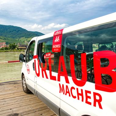 Weißer Radweg-Reisen Urlaubmacher Bus mit roter Beschriftung und Fahrrädern im Inneren, auf Holzsteg am Fluss mit Hügeln und Dorf im Hintergrund.