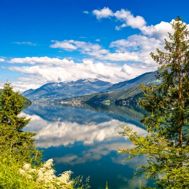 Millstätter See mit ruhigem türkisfarbenem Wasser, das Berge und Wolken spiegelt. Grüne Bäume rahmen die Aussicht, Berge erheben sich im Hintergrund.
