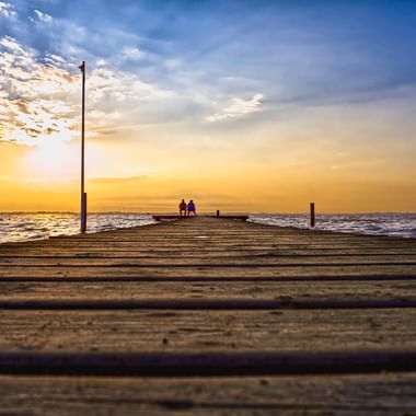 Holzsteg, der bei Sonnenuntergang in die Nordsee bei Dangast führt. Zwei Personen sitzen am Ende des Stegs, als Silhouetten vor goldenem Himmel.
