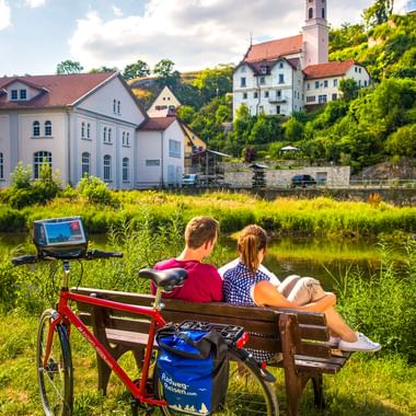 Zwei Radfahrer auf einer Bank an der Donau mit rotem Fahrrad, Blick auf Rechtenstein mit Kirchturm und weißen Gebäuden.