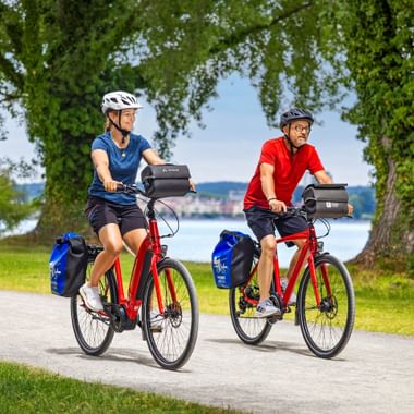 Zwei Radfahrer auf roten Tourenrädern mit Packtaschen fahren durch den Seeburgpark in Kreuzlingen. Bodensee und Konstanz im Hintergrund.
