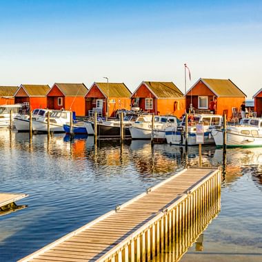 Hafen Rødvig mit Holzsteg, weißen Booten an Anlegestellen und orange-roten Bootshäusern unter klarem blauen Himmel. Ruhiges Wasser spiegelt die Szene.