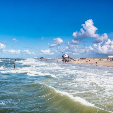 Breiter Sandstrand in St. Peter-Ording mit Wellen der Nordsee. Hölzerne Pfahlbauten stehen am Strand unter blauem Himmel mit Wolken.