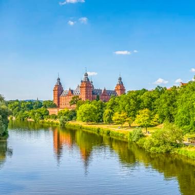 Blick auf Schloss Johannisburg mit seinen roten Sandsteintürmen über grünen Bäumen entlang des Mains in Aschaffenburg unter blauem Himmel.