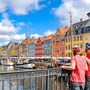 Zwei Radfahrer mit Helmen betrachten bunte historische Gebäude entlang des Nyhavn-Hafens in Kopenhagen mit Booten im Kanal.