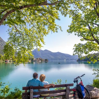 Zwei Radfahrer sitzen auf einer Holzbank unter grünen Bäumen und blicken auf den türkisfarbenen Walchensee mit Bergen im Hintergrund. Ein Fahrrad steht daneben.