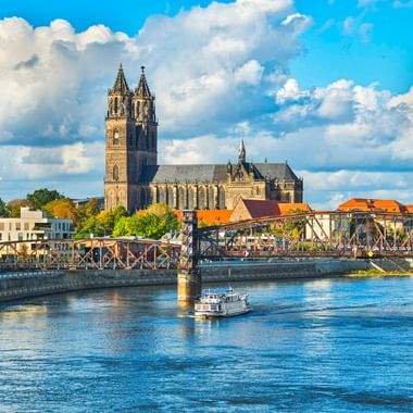 Magdeburger Dom mit Doppeltürmen über der Elbe. Eine Stahlbrücke überquert den Fluss, darunter fährt ein Boot an einem sonnigen Tag.