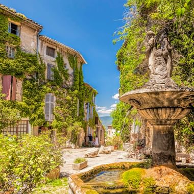 Steinbrunnen mit Statue auf einem sonnigen Platz in Saignon, Provence. Efeubedeckte Gebäude und üppiges Grün umgeben den verwitterten Brunnen.