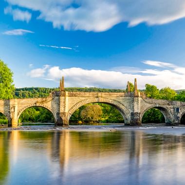 Historische Steinbrücke mit fünf Bögen über einem Fluss in Aberfeldy. Grüne Bäume säumen beide Ufer unter blauem Himmel mit weißen Wolken.