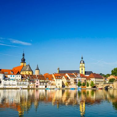 Kitzinger Mainufer mit bunten historischen Gebäuden und Kirchtürmen, die sich im Main spiegeln, unter klarem blauen Himmel in Franken.