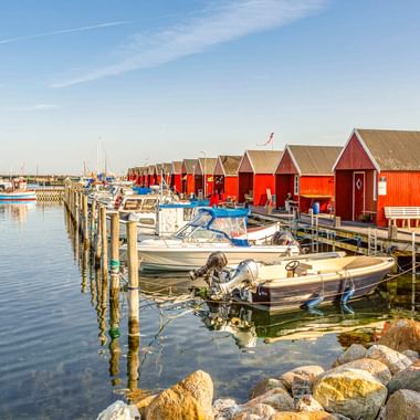 Hafen Rødvig mit roten Holzbootshäusern auf Holzstegen, kleinen Booten im ruhigen Wasser und Felsen entlang des Ufers unter blauem Himmel.