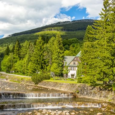 Kaskadierender Bach mit Steinstufen in Spindlermühle, weißes Haus zwischen grünen Bäumen, bewaldeter Berg unter blauem Himmel mit Wolken.