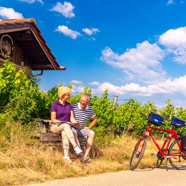 Zwei Radfahrer sitzen auf Stufen neben einer Weinberghütte in Baden-Württemberg. Ein rotes Tourenrad mit blauen Taschen steht auf dem Weg. Grüne Reben und blauer Himmel sichtbar.