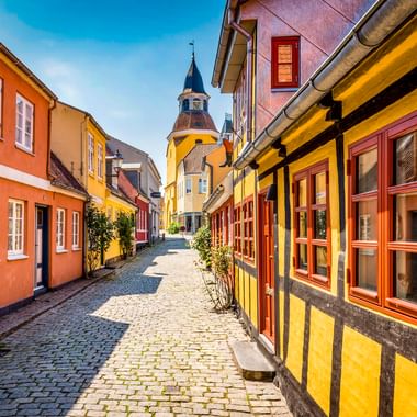 Kopfsteinpflasterstraße in der Faaborg Altstadt mit bunten Fachwerkhäusern in Gelb, Orange und Rot. Ein Kirchturm erhebt sich im Hintergrund.