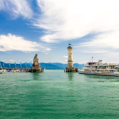 Lindauer Hafeneinfahrt mit Löwenstatue und Leuchtturm am Bodensee. Ein Passagierschiff fährt vorbei, Segelboote im Hintergrund, Berge sichtbar.