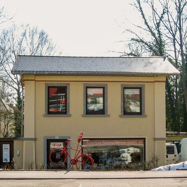 Gelbes zweistöckiges Gebäude mit grauem Dach und drei Fenstern. Rotes Fahrrad davor. Schild mit 'Bodensee-Radweg Service GmbH'.