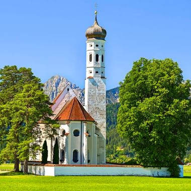 St. Coloman Kirche mit weißem Turm und Zwiebelkuppel steht auf grüner Wiese bei Schwangau, eingerahmt von Bäumen mit Alpenbergen im Hintergrund.