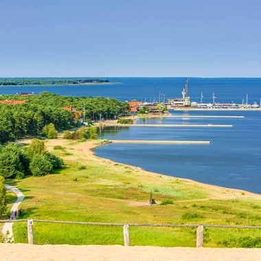 Panoramablick von der Parnidis-Düne in Nidden mit Sandstrand, blauem Ostseewasser, grünem Wald und Yachthafen unter klarem Himmel.