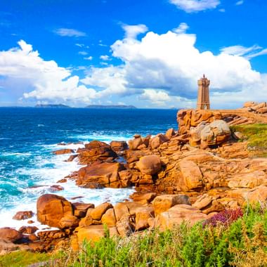 Leuchtturm Phare de Ploumanach auf rosa Granitfelsen am blauen Atlantik unter bewölktem Himmel, mit grüner Küstenvegetation.