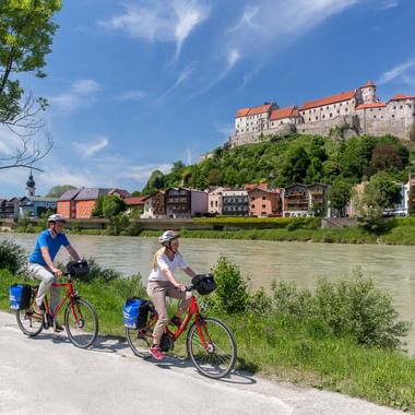 Zwei Radfahrer mit roten Rädern und blauen Taschen fahren auf einem Flussweg mit der Burg Burghausen auf einem Hügel und bunten Stadthäusern darunter.