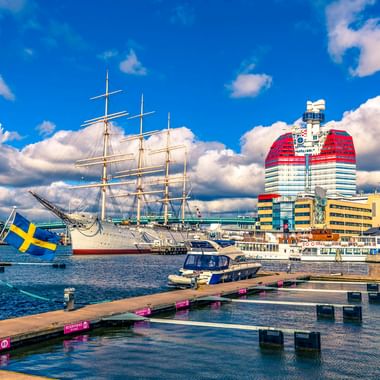 Göteborger Hafen mit weißem Segelschiff, Holzstegen mit schwedischer Flagge und modernem rot-weißem Gebäude unter blauem Himmel.