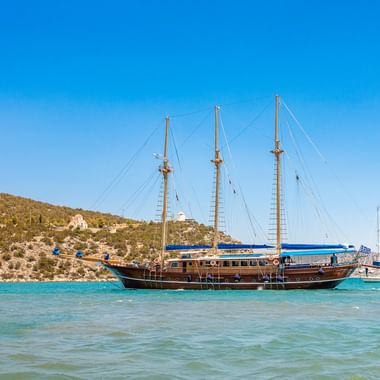 Dreimaster-Segelschiff Ermioni Thalassa mit blauem Zierstreifen ankert in türkisfarbenem Mittelmeer vor felsiger Küste unter blauem Himmel.