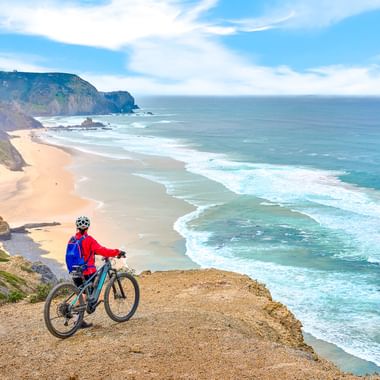 Radfahrerin in roter Jacke auf Mountainbike an Klippenrand mit Blick auf Strand und türkisfarbenen Atlantik bei Sagres, Portugal.