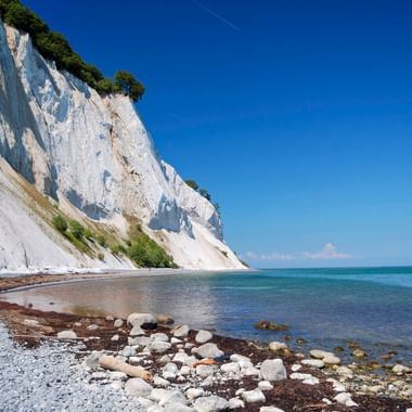 Dramatische weiße Kreidefelsen von Møns Klint, die sich von einem Kieselstrand entlang der türkisfarbenen Ostsee unter blauem Himmel erheben.