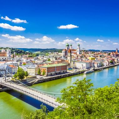 Panoramablick auf die Passauer Altstadt mit historischen Gebäuden und Kirchtürmen entlang der Donau unter blauem Himmel mit weißen Wolken.
