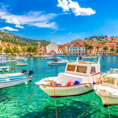Bunte Boote im kristallklaren türkisfarbenen Wasser des Hafens von Hvar, mit mediterranen Gebäuden, Palmen und grünen Hügeln unter blauem Himmel.