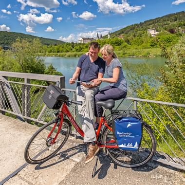 Paar auf rotem Fahrrad mit blauen Taschen betrachtet Karte auf Brücke über die Altmühl bei Riedenburg, mit grünen Hügeln und Kirchtürmen.