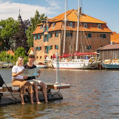 Zwei Radfahrer sitzen auf einem schwimmenden Steg im Hafen Glückstadt mit den Füßen im Wasser, Fahrräder neben sich. Historische Gebäude und Segelboote im Hintergrund.