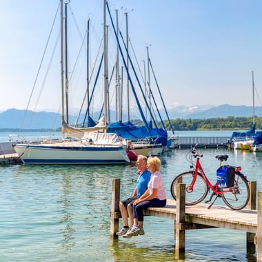 Zwei Radfahrer sitzen auf einem Holzsteg am Starnberger See mit einem roten Fahrrad neben sich. Segelboote im Hafen, Berge im Hintergrund.