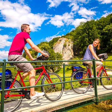 Zwei Radfahrer auf einer Metallbrücke über die Donau bei Schelklingen. Weiße Kalksteinfelsen und grüner Wald im Hintergrund.