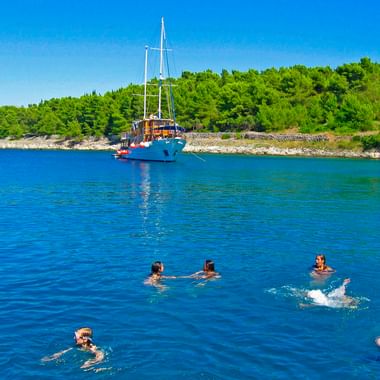 Six people swimming in clear blue water near the MS Mirabela sailing ship anchored in a bay with green forested coastline under blue sky.