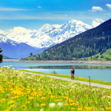 Radfahrer auf Weg am Reschensee mit Wildblumenwiese, schneebedeckten Alpen im Hintergrund unter blauem Himmel in Südtirol.