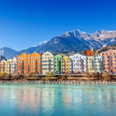 Row of colorful historic buildings along the turquoise Inn River in Innsbruck, with snow-capped Tyrolean Alps in the background under clear blue sky.