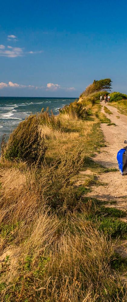 Zwei Radfahrer auf roten Rädern auf einem Sandweg entlang der Steilküste bei Nienhagen, Ostsee. Strand mit Wellen und Dünengras sichtbar.