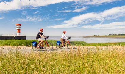 Zwei Radfahrer mit roten Helmen und blauen Taschen fahren entlang eines Weges am Wasser in Brunsbüttel. Ein rot-weißer Leuchtturm steht am Wasser.