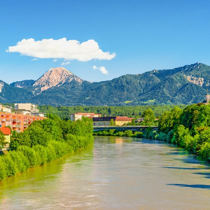 The Drau River flowing through Villach, Carinthia, with green vegetation along the banks, a bridge, and dramatic mountain peaks in the background.