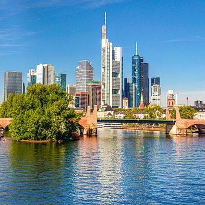 Frankfurter Skyline mit modernen Wolkenkratzern und historischen Kirchtürmen entlang des Mains. Steinbrücke überquert das Wasser unter blauem Himmel.
